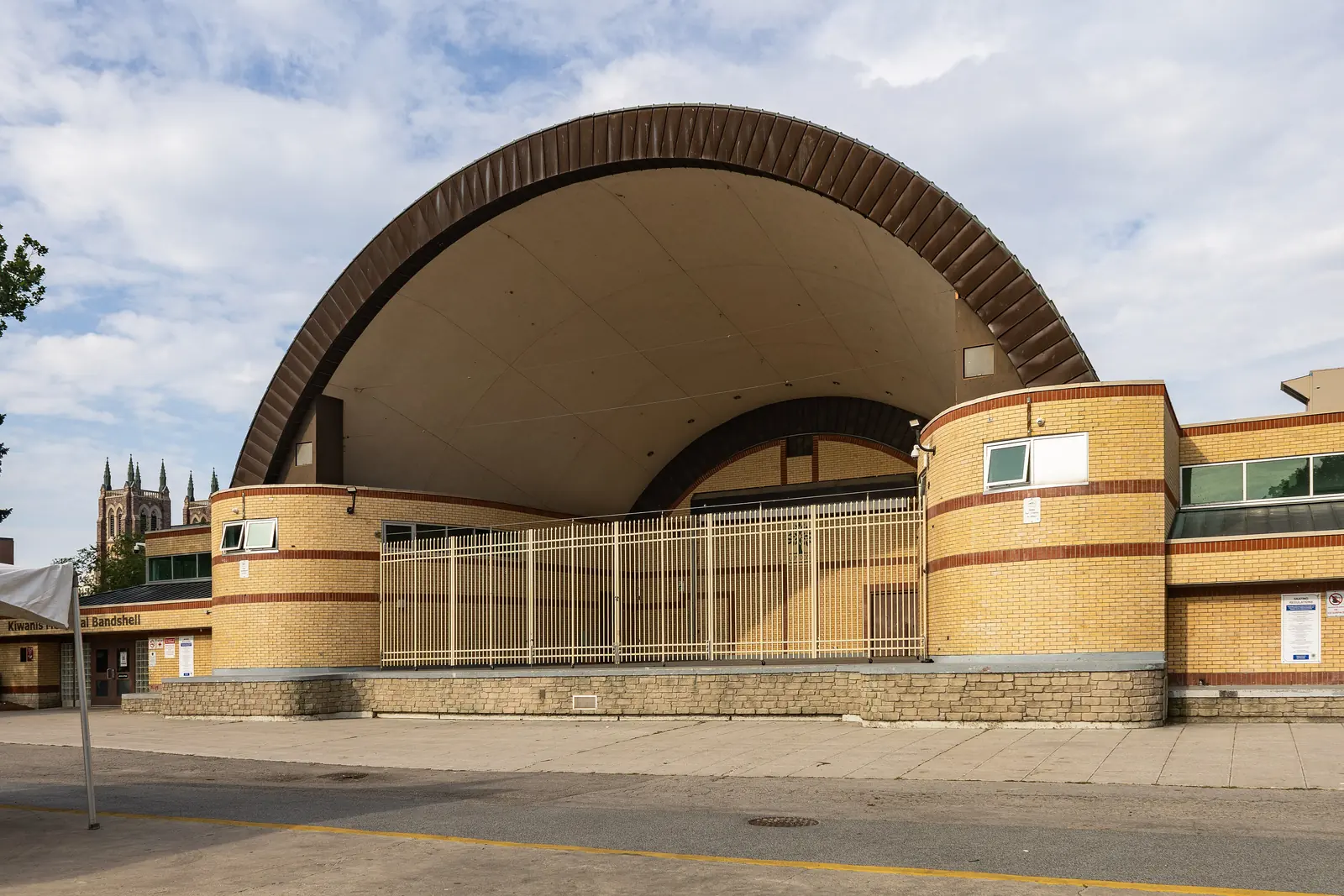 The Kiwanis Memorial Bandshell in Victoria Park, London, Ontario, a brick and concrete shell with a large arch, and church spires behind the trees to the left.